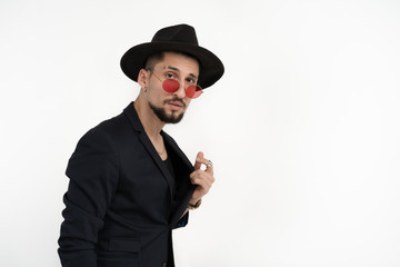 Pleased business man with beard in black suit and hat, in red sunglasses, holding jacket and looking at the camera isolated over white background