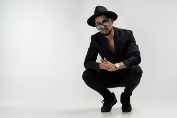 Handsome bearded man in black suit and trendy hat in black sunglasses sat down and rubs his hands, looking at the camera, isolated over white background