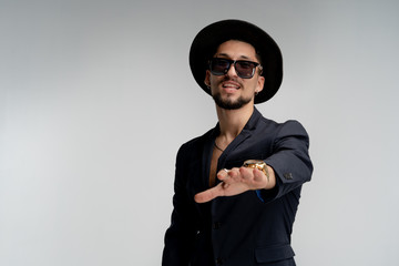 Obraz na płótnie Canvas Happy attractive young bearded man in stylish black suit and hat in black sunglasses, posing with outstretched hand isolated over white background