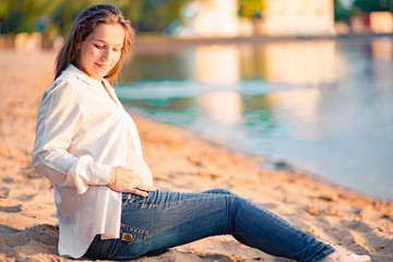Beautiful pregnant woman sitting by the river at sunset