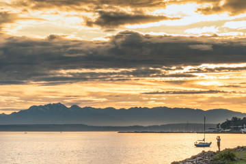 Silhouetted photographer at sunset on waters edge with sailboat.