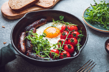 Breakfast time, fried egg with sausages and cherry tomatoes in a black iron pan, served microgreens.