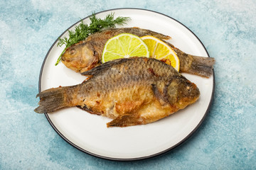 Fried carp fish on white plate with lemon and lime slices, top view. Ready meal. Blue background