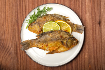 Fried carp fish on white plate with lemon and lime slices, top view. Ready meal. Wooden background