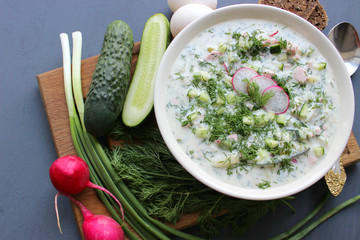 Okroshka is a traditional Russian summer cold soup in a ceramic bowl. Ingredients potatoes, radishes, cucumbers, dill, eggs, kefir (yogurt), sausage. Summer yogurt cold soup on a wooden table. Selecti