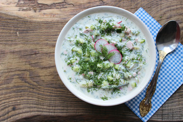 Okroshka is a traditional Russian summer cold soup in a ceramic bowl. Ingredients potatoes, radishes, cucumbers, dill, eggs, kefir (yogurt), sausage. Summer yogurt cold soup on a wooden table. Selecti