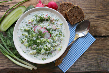 Okroshka is a traditional Russian summer cold soup in a ceramic bowl. Ingredients potatoes, radishes, cucumbers, dill, eggs, kefir (yogurt), sausage. Summer yogurt cold soup on a wooden table. Selecti