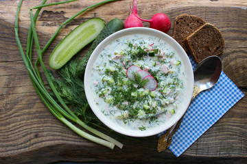 Okroshka is a traditional Russian summer cold soup in a ceramic bowl. Ingredients potatoes, radishes, cucumbers, dill, eggs, kefir (yogurt), sausage. Summer yogurt cold soup on a wooden table. Selecti
