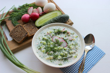 Okroshka is a traditional Russian summer cold soup in a ceramic bowl. Ingredients potatoes, radishes, cucumbers, dill, eggs, kefir (yogurt), sausage. Summer yogurt cold soup on a wooden table. Selecti