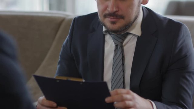Tilt up of HR manager wearing official style clothes sitting on couch, listening to employee sitting in front of him and studying his CV