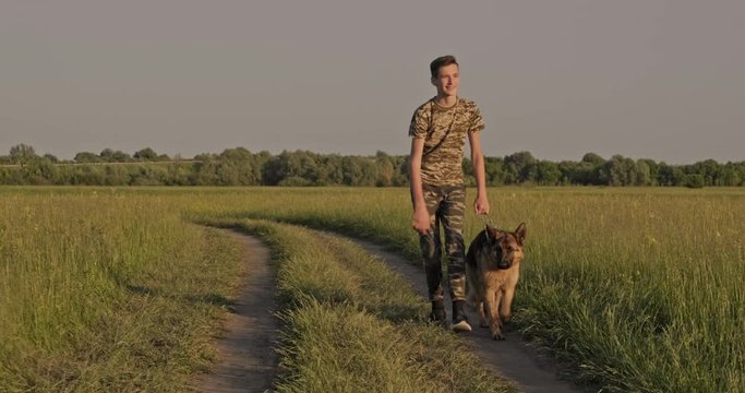 Teenager Is Walking  With A Dog In Nature. 15 Year Old Boy Strolls With A Dog Breed German Shepherd. Teen Spends Time On The Field With Dog. Caucasian Guy  With His Pet In The Meadow. Real Time