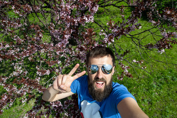 Excited guy doing sefie with peace gesture outdoors near bloossom cherry tree