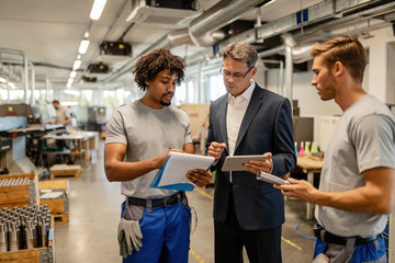 African American worker and his manager going through reports and using touchpad in steel factory.