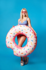 Full-length photo of young woman in bathing suit with inflatable donut on empty blue background