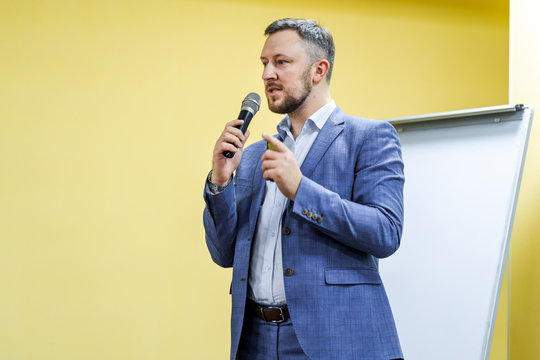 Bearded Businessman Is Speaking To The Mic On The Light Hall Background. Handsome Entepreneur Leading The Meeting During Important Symposium Indoors.