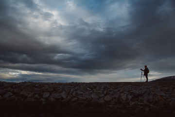 Silhouette of a photographer or traveler with tripod standing on stone. Background of a dramatic sky. Hazardous working conditions and human silhouette