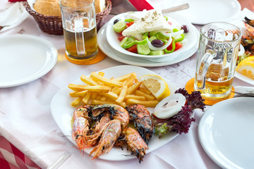 Plate with fresh greek salads, shrimp, potato fried, beers and bread, olive oil on a table. Traditional Greek dish.