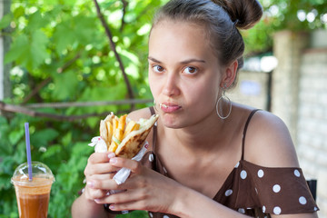 beautiful young girl with pleasure eating a hamburger (Greek gyros), girl in a dress sitting in a street cafe, eating a hamburger
