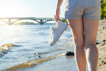 Fototapeta na wymiar girl walks along the beach with running shoes in her hands