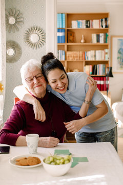Smiling young female volunteer embracing retired senior woman sitting at table in nursing home
