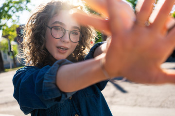 Outdoor close up portrait of young beautiful stylish happy smiling curly girl wearing sunglasses, posing in street. Sunny day light. Summer fashion concept. Copy, empty space for text