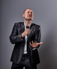 Smiling emotional moving happy performer man presenting the show holding microphone in hand and showing the finger the choosing sign on grey color background. Closeup