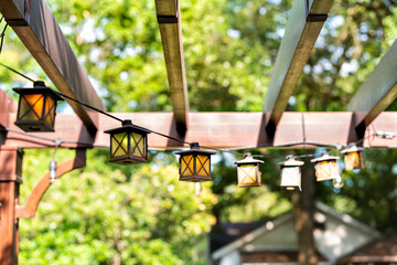 Closeup of patio outdoor spring garden in backyard of home with lantern lamps lights hanging from pergola canopy wooden gazebo and plants - obrazy, fototapety, plakaty