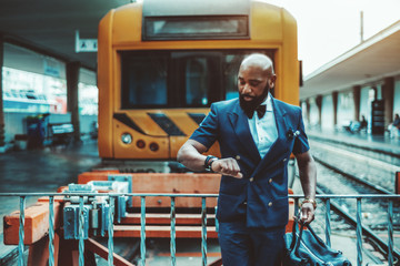 Stylish bald bearded African man entrepreneur in a vest is checking the departure time for his business trip on his watch while standing with a travel bag in front of a high-speed train on a platform