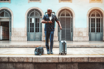 A handsome bald bearded African male entrepreneur in a fashion vest and glasses is waiting for his train and looking at his watch on the railway platform to go on a business trip with two travel bags - Powered by Adobe