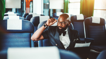 Portrait of an adult bald fashion African businessman with beard looking out the window adjusting his eyeglasses while sitting indoors of the first-class car of a high-speed train