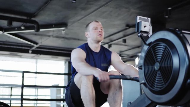 Strong middle aged man doing exercise with rope pull machine and resting after set in gym, tracking left medium shot