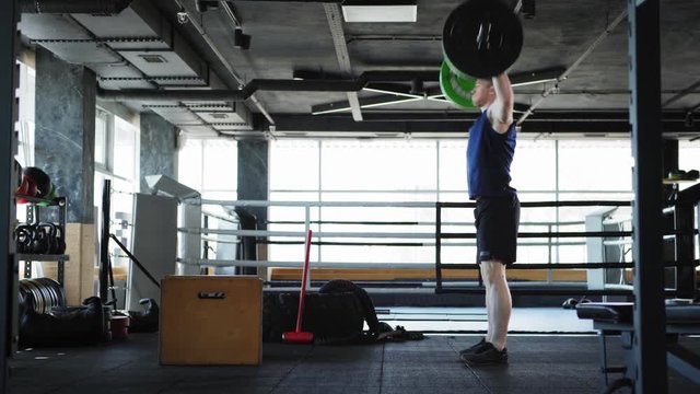 Side view of athletic middle aged man doing overhead barbell lifts and box jumps exercise. Man recovering after workout set in gym