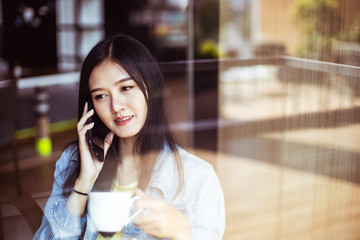Asian woman with beautiful smile hold mobile phone during rest in coffee shop.Attractive woman drinking coffee.Reflection glass window.