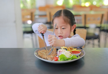 Asian child girl eating Pork steak and vegetable salad on the table with holding knife and fork....