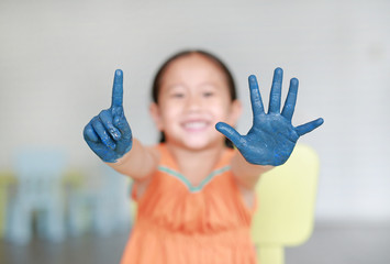 Happy little Asian girl with her blue hands painted showing one and five fingers in kids room.