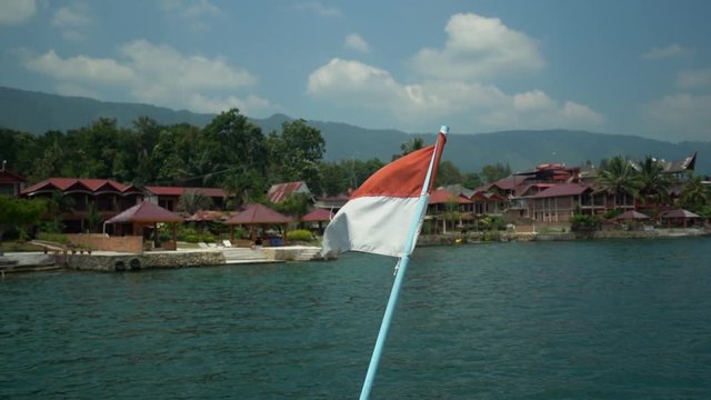 Slow motion of the Indonesia flag blowing in the wind on a ferry boat crossing Lake Toba with traditional resorts in the background in North Sumatra.