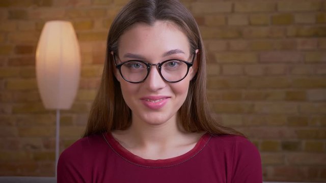 Portrait of young and beautiful brunette female in eye glasses smiles prettily into camera in home.