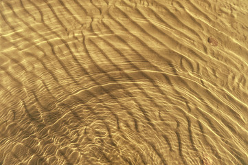 Waves on a yellow sandy bottom intersect with waves on the surface of the water. The texture of sand and water.