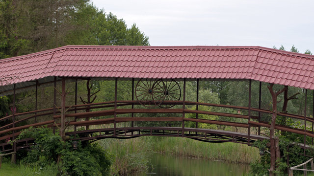 A Wooden Bridge With Two Wooden Cart Wheels And A Railing With A Dark Red Metal Tile Roof Over A Small River On A Background Of Green Trees And Bushes