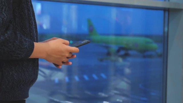 daughter traveler with a backpack airplane flights airport concept. girl teenager with a smartphone looks out the window at the airport area airplanes and bus at a distance. teen girl looks at the