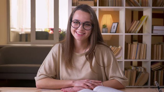 Closeup portrait of young attractive caucasian female student in glasses looking at camera smiling happily indoors in the college library