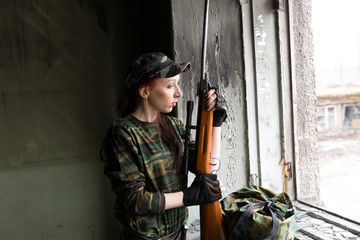 A thin red-haired girl stands at the broken window with a rifle. The girl in green camouflage with...
