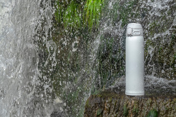 Naklejka na ściany i meble White thermos of tea or coffee on stone near waterfall with splashes of water drops. Adventure hiking tourism concept.