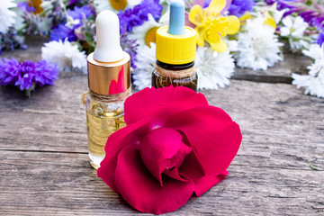 essential oil with a rose in a glass bottle near wildflowers on wooden background.