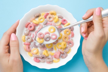 Hands holding white bowl and spoon on pastel blue table. Woman eating colorful, bright cereal rings...