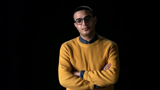 Medium shot of young Arab man wearing yellow sweater and eyeglasses looking at camera and crossing arms on his chest
