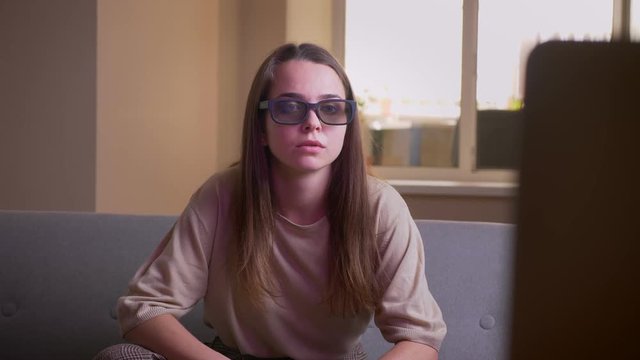 Closeup portrait of young attractive caucasian female watching a 3D horror movie on TV getting a jumpscare sitting on the couch indoors in the apartment