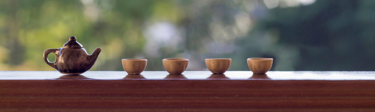Little brown ceramic teapot and cups on wooden stand, green garden background. Close-up, copy space, horizontal layout.