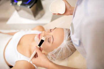 Woman closing eyes while dermatologist putting mask with brush