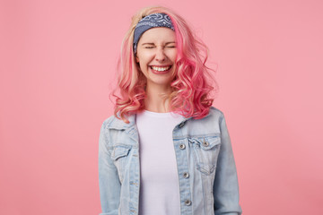 Happy cute smiling lady with pink hair and tattooed hands, standing over pink background with closed eyes, broadly smiling, wearing a white t-shirt and denim jacket.